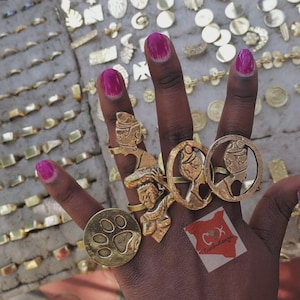 May include: A hand wearing several gold rings with various designs, including a paw print, a profile, and an Egyptian figure. The background displays a variety of gold jewelry, including rings and earrings. The nails are painted a bright pink.