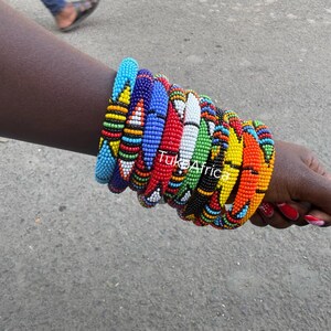 Può includere: Una collezione di braccialetti di perline colorate, tra cui blu, rosso, giallo e verde. I braccialetti sono impilati su un polso, mostrando motivi e disegni intricati. L'immagine è stata scattata all'aperto.