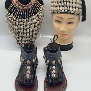 May include: A collection of handcrafted African accessories. It includes a beaded necklace with cowrie shells, a hat decorated with cowrie shells and a black tassel, and black sandals with cowrie shell accents. The sandals have brown leather soles.