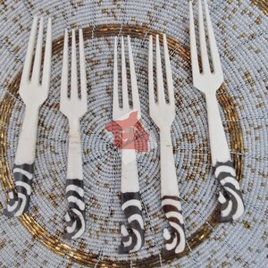 May include: A set of five ivory-colored forks with a distinctive design. The handles feature a black and white zebra stripe pattern. The forks are arranged on a beaded placemat with gold and silver accents, creating a decorative dining set.