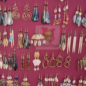 May include: A collection of various handcrafted earrings displayed on a burgundy surface. The earrings feature diverse designs, including gold wire spirals, teardrop shapes, and those adorned with shells, beads, and bone. The color palette includes gold, black, white, and turquoise.