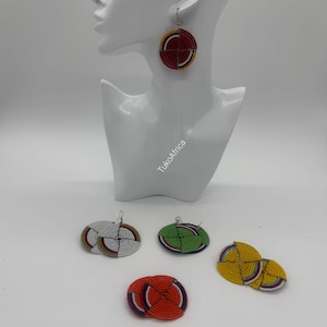 Puede incluir: Pendientes hechos a mano con cuentas de varios colores, incluyendo rojo, amarillo, verde y blanco. Los pendientes son circulares con un diseño geométrico. Un par se muestra en una cabeza de maniquí blanca. La marca "TukoAfrica" es visible.
