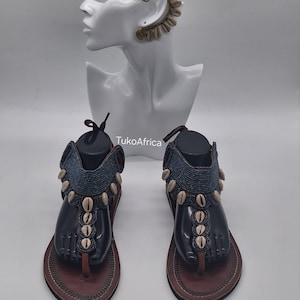 May include: A pair of brown leather thong sandals featuring dark blue beaded straps adorned with white cowrie shells, displayed alongside a white mannequin head wearing a matching ear cuff with a spiral wire design and cowrie shells.