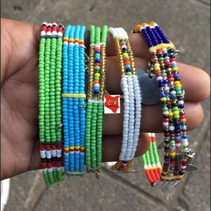May include: Several beaded bracelets in various colours are displayed. The bracelets feature green, blue, white, and multicoloured beads. Some have metal accents and tags. The bracelets are likely handmade and showcase a vibrant, ethnic design.