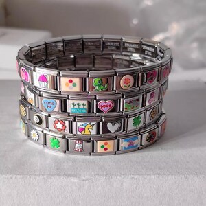 Puede incluir: Pila de pulseras de abalorios de acero inoxidable con coloridos abalorios de esmalte. Los abalorios incluyen una magdalena, un dinosaurio verde, un corazón con la palabra "love", un trébol de cuatro hojas y la palabra "Arizona".