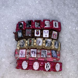 Op de afbeelding: Gestapelde armbanden met rechthoekige kralen met verschillende afbeeldingen. De armbanden zijn in roze, bruin en beige. De afbeeldingen op de kralen omvatten portretten en illustraties. De armbanden worden gepresenteerd op een wit, getextureerd oppervlak.