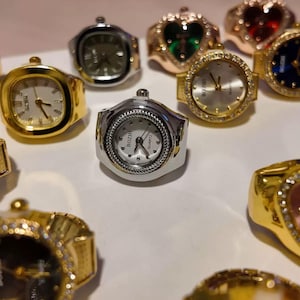 Puede incluir: Una colección de relojes de anillo en varios estilos. Los relojes tienen esferas redondas o rectangulares con monturas doradas o plateadas. Algunos tienen elementos decorativos como piedras preciosas o biseles texturizados. Las esferas muestran la hora con manecillas.
