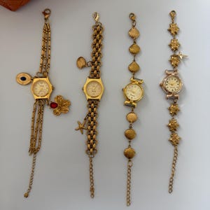 May include: Four gold-toned bracelet watches with different designs. One has a chain with a flower and a charm, another has a chain with a starfish charm, and the other two have seashell and star accents. The watches have gold or silver watch faces.