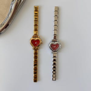 May include: Two heart-shaped face watches with matching metal bands. One watch is gold-toned with a red face, the other is silver-toned with a red face. The watches are displayed on a white surface.