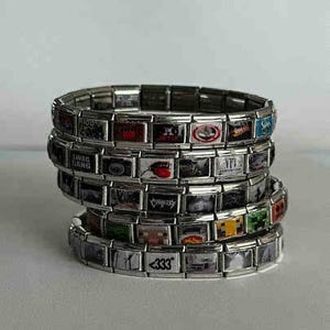 May include: Stack of silver-toned charm bracelets. Each bracelet is composed of rectangular links, some of which feature colorful graphics and text. The bracelets are stacked on top of each other.