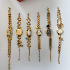 Könnte beinhalten: Eine Sammlung von sechs goldfarbenen Vintage-Armbanduhren mit verschiedenen Zifferblattformen und Armbändern im Kettenstil. Einige Uhren verfügen über dekorative Anhänger und hängende Ketten. Die Uhren werden auf einer weißen Oberfläche präsentiert, mit einem braunen Lederartikel im Hintergrund.