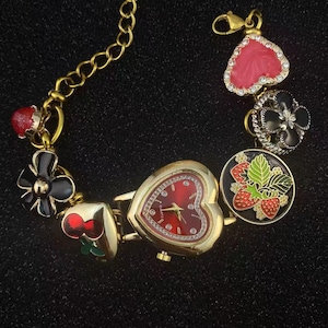 Peut inclure: Montre-bracelet dorée avec un cadran rouge en forme de cœur et des breloques décoratives. Les breloques comprennent un cœur rouge, une fleur noire, une cerise et un motif de fraise. La montre a une chaîne et un fermoir dorés.