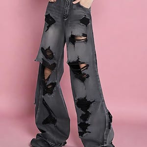 Pu&ograve; includere: Jeans grigi a gamba larga strappati con grandi lacerazioni. I jeans hanno una vita alta e una cintura nera con fibbia argentata. I jeans sono abbinati a scarpe nere. Lo sfondo &egrave; rosa.