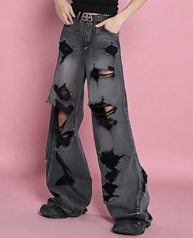Pu&ograve; includere: Jeans grigi a gamba larga strappati con grandi lacerazioni. I jeans hanno una vita alta e una cintura nera con fibbia argentata. I jeans sono abbinati a scarpe nere. Lo sfondo &egrave; rosa.