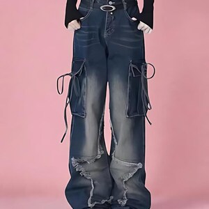 May include: Dark wash denim cargo pants with distressed details. The pants feature large pockets with drawstrings and a wide-leg fit. The denim has a faded, worn look with a light wash effect on the knees and thighs. The pants are paired with black sneakers.
