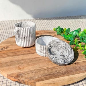 Peut inclure: Un ensemble de trois contenants décoratifs en béton avec un motif marbré. L'ensemble comprend un cylindre haut et strié, un petit bol et un couvercle ovale. Les articles sont disposés sur une planche de service en bois.