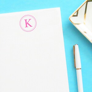 May include: A white notepad with a pink letter "K" in a circle of pink dots. A white pen and a white and gold decorative dish are on a blue background.