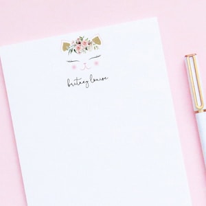 May include: White notepad with a floral cat illustration and the name "brittany louise" printed on it. A white pen with gold accents is lying next to the notepad on a pink surface.