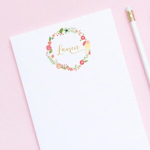 Pu&ograve; includere: Un blocco note bianco con un design a corona di fiori e il nome "Lauren" scritto in caratteri dorati. Il blocco note &egrave; su una superficie rosa con una matita bianca.