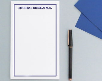 May include: A white notepad with a blue border and the text "MICHEAL EINMAN M.D." at the top. A black pen is lying on a gray surface next to the notepad.
