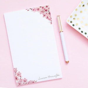 May include: A white notepad with a pink floral design on the top and bottom corners. The notepad has the name "Lauren Herrington" written on the bottom right corner. A white pen with a gold top is lying on a pink surface next to the notepad.