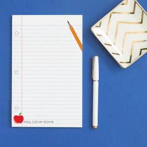 May include: A white lined notepad with a red apple and the text "Miss Silverstone" on the bottom left corner. A yellow pencil is on the notepad and a white pen is on a blue surface.