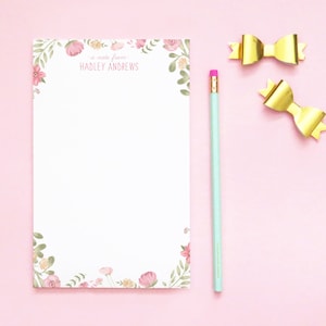 May include: A white notepad with floral corner designs, the text "a note from HADLEY ANDREWS", a light blue pencil, and two gold bow-shaped paper clips. The notepad has pink and green floral accents.