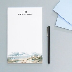 May include: A white notepad with a watercolor beach scene at the bottom. The notepad has the initials "LS" and the name "LAUREN SMITHSTONE" printed at the top. A black pen and two light blue envelopes are next to the notepad.