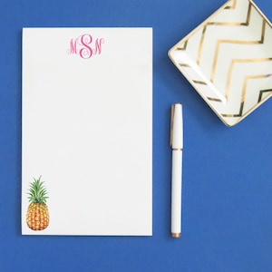 May include: A white notepad with a pink monogram "MSN" at the top and a pineapple illustration at the bottom. A white pen and a gold and white decorative dish are also in the image.