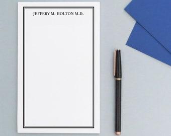 May include: A white notepad with a black border and the text "JEFFERY M. HOLTON M.D." at the top. A black pen with a gold top is lying on a light blue surface.