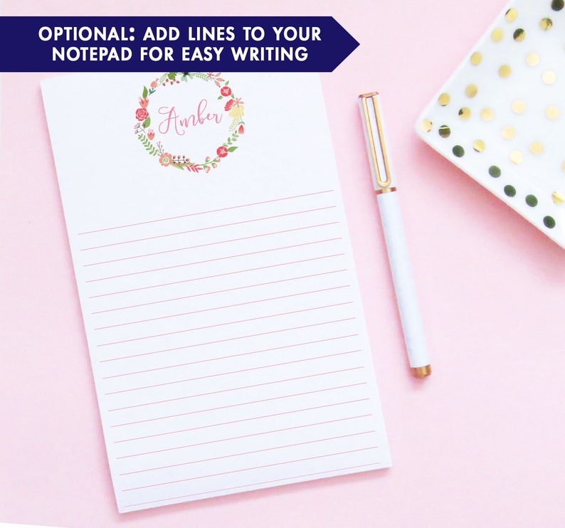 Pu&ograve; includere: Un blocco note bianco con carta rigata rosa e una ghirlanda di fiori con il nome "Amber" in lettere rosa. Il blocco note &egrave; su uno sfondo rosa con un piatto bianco con pois dorati.