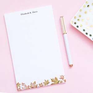 May include: A white notepad with a personalized name, "Elizabeth R. Davis", printed at the top. The notepad has a border of brown and orange autumn leaves at the bottom. A white pen with gold accents is lying next to the notepad.