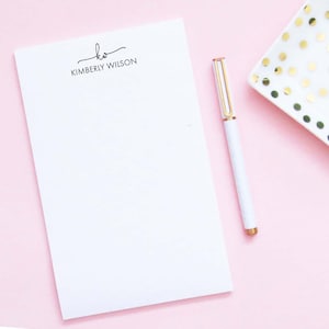 May include: A white notepad with a black handwritten signature and the name "KIMBERLY WILSON" printed below. A white pen with gold accents is lying on a pink surface.