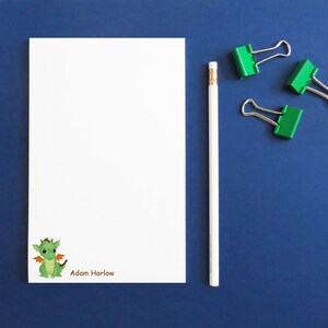 May include: White notepad with a green dragon illustration and the name "Adam Harlow" printed in brown. A white pencil and three green binder clips are on a blue surface.