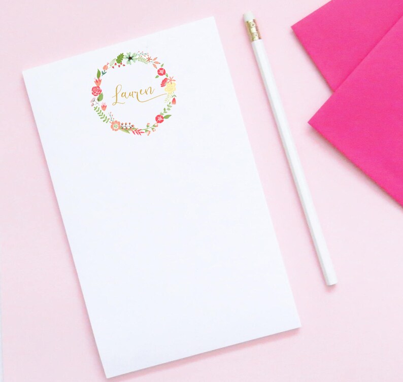 Pu&ograve; includere: Un blocco note bianco con un design a corona di fiori e il nome "Lauren" scritto in caratteri dorati. Il blocco note &egrave; su una superficie rosa con una matita bianca.