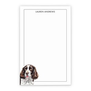 May include: A white rectangular notepad featuring a thin black border and the name LAUREN ANDREWS printed in black at the top. The bottom left corner displays a watercolor illustration of a brown and white spaniel dog with long ears.