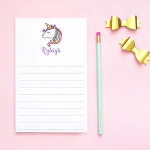 May include: A white notepad with a unicorn illustration and the name "Ryleigh" printed in purple. The notepad has lined paper and is on a pink background with a gold bow and a blue pencil.