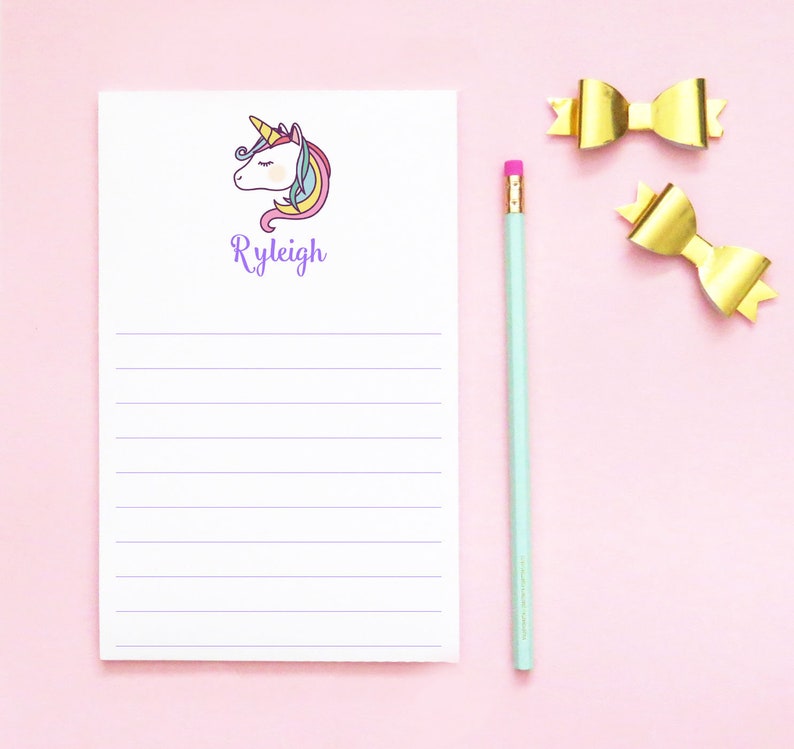 May include: A white notepad with a unicorn illustration and the name "Ryleigh" printed in purple. The notepad has lined paper and is on a pink background with a gold bow and a blue pencil.