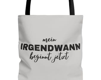 May include: A cream-coloured tote bag with black handles. The bag displays the German phrase "mein IRGENDWANN beginnt jetzt" in black. The word "mein" is in a smaller font above "IRGENDWANN", and "beginnt jetzt" is in a script font below.