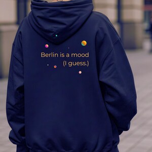 Peut inclure: Sweat à capuche bleu marine avec l'inscription "Berlin is a mood (I guess.)" en or. Le design comprend des graphiques de points colorés. Le sweat à capuche a une capuche et une poche avant.