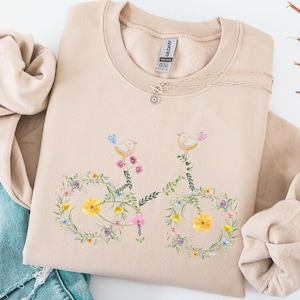 Peut inclure: Sweat-shirt beige avec un motif de vélo floral. Le vélo est composé de fleurs et de feuilles colorées, avec deux oiseaux perchés au sommet. Un délicat collier argenté avec un petit pendentif est visible. Le sweat-shirt est associé à un jean bleu.