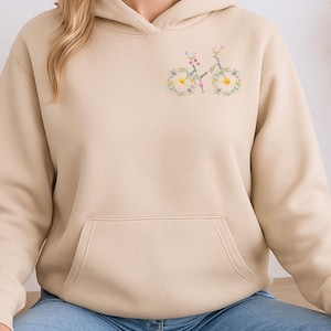 Peut inclure: Un sweat à capuche beige avec une poche avant et un motif de vélo floral sur la poitrine. Les roues du vélo sont faites de fleurs jaunes et le cadre est vert. Le sweat à capuche est en matière douce.
