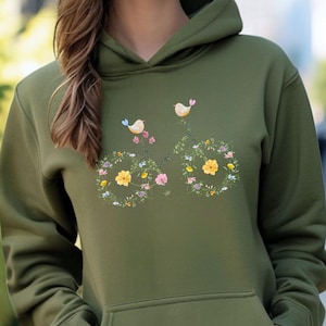 Peut inclure: Un sweat à capuche vert olive avec un motif de vélo floral. Le motif comprend des fleurs jaunes, roses et bleues, et deux petits oiseaux. Le sweat à capuche a une poche avant et une capuche à cordon.