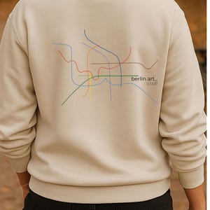 May include: Cream-coloured sweatshirt with a colourful abstract map design on the back, including the text "berlin art_ U1/09". The sweatshirt has a crew neck and is a neutral colour.
