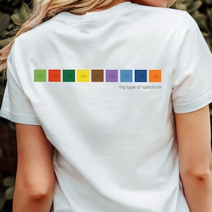 Puede incluir: Camiseta blanca con un diseño de espectro colorido en la espalda. El diseño presenta nueve cuadrados de colores con el texto "U1" a "U9" y la frase "my type of spectrum" debajo.