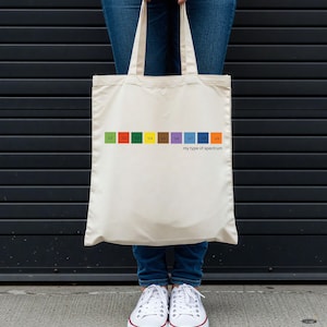 Puede incluir: Un bolso tote de color crema con un diseño de espectro colorido y el texto "my type of spectrum". El bolso lo sostiene una persona que lleva vaqueros azules y zapatillas blancas.