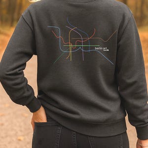 May include: Charcoal grey sweatshirt with a colourful, abstract line design, similar to a city map. The text "berlin art. club" is printed on the right side of the design. The sweatshirt has a crew neck.