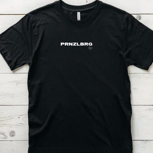 Könnte beinhalten: Schwarzes T-Shirt mit dem weißen Text "PRNZLBRG" und "ofo" auf der Vorderseite. Das Shirt besteht aus einem weichen Material und hat einen Rundhalsausschnitt. Das T-Shirt wird auf einer weißen Holzoberfläche präsentiert.