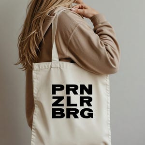 Puede incluir: Bolso tote de color crema con el texto "PRN ZLR BRG" en letras negras en negrita. El bolso tiene un diseño sencillo con correas largas para llevar al hombro. El bolso está hecho de un material resistente.