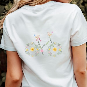 May include: White t-shirt featuring a bicycle design made of flowers and leaves. The bicycle has two birds perched on top. The design includes yellow, pink, and green floral elements, creating a whimsical and nature-inspired aesthetic.